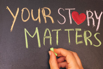 your story matters phrase handwritten on blackboard with heart symbol instead of O, hand holding...