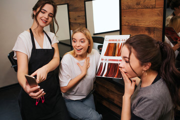 Surprised young women looking at phone while hairdresser holding it in hand. Hairstylist proposing fashion hair coloring in salon