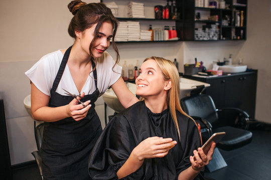 Professional hairdresser with scissors and beautiful female customer deciding what haircut to do while looking at phone. Changing haircut in beauty salon
