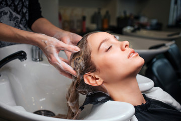 Beautiful young woman sitting near sink while hairdresser washing her hair in beauty salon. Hair stylist at work