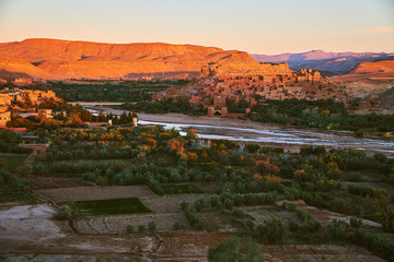 Early morning sun hitting the top of historic heritage Ait Ben Haddou with green oasis in desert surrounded by dry mountains in Morocco Africa