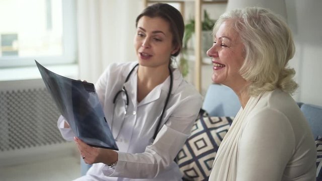 Qualified and experienced specialist with stethoscope on neck looking and discuss X-rays picture with old retired lady. They sitting inside bright living room and smiling