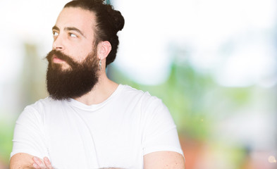 Young hipster man with long hair and beard wearing casual white t-shirt smiling looking to the side with arms crossed convinced and confident