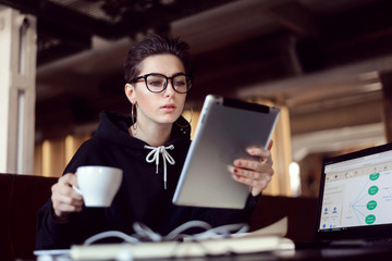 Portrait of shorthair serious female student wearing glasses sitting with the tablet during break in cafe