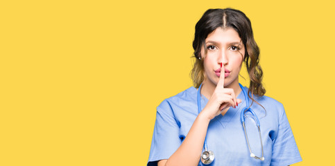 Young adult doctor woman wearing medical uniform asking to be quiet with finger on lips. Silence...