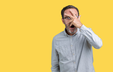 Handsome middle age elegant senior man wearing glasses over isolated background peeking in shock covering face and eyes with hand, looking through fingers with embarrassed expression.