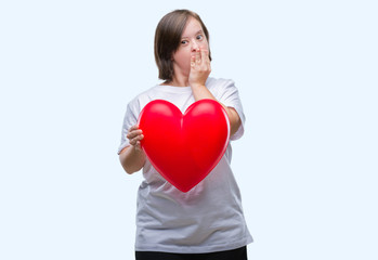 Young adult woman with down syndrome holding red heart over isolated background cover mouth with...