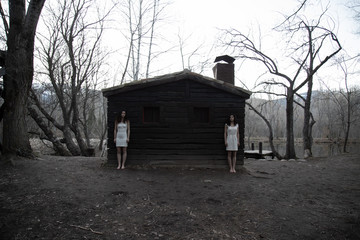Young twin sisters in white dresses standing near abandoned wood house in mysterious forest