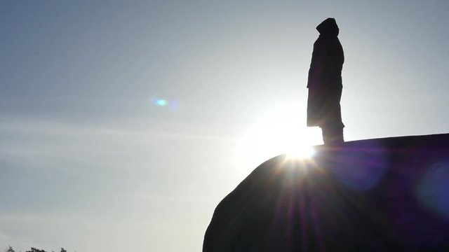standing monk in hoodie praying at the edge of rock silhouette