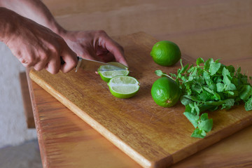 close up of man's hands with  knife cut lime, lime and mint on a wooden cutting board