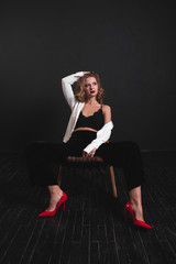 Fototapeta na wymiar Beautiful, sexy and fashionable blonde girl with red lips in white shirt, black bra and black leggings sits on a wooden chair on black background
