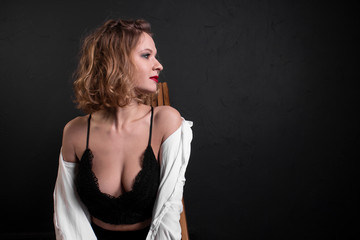 Fototapeta premium Beautiful, sexy and fashionable blonde girl with red lips in white shirt, black bra and black leggings sits on a wooden chair on black background