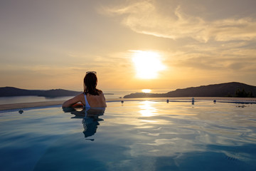 Woman in infinity swimming pool at golden sunset. Girl admiring sunset from the pool. Summer vacation holiday