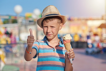Cute European boy with the hat on his head and in striped t-shirt eating ice-cream in the Luna park. He is making like gesture.