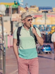 European man in the sunhat is walking around the Luna Park and eating tasty ice-cream.