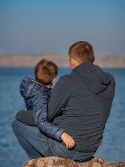 Cute European boy and his dad are sitting on the stone at the bank of the lake. They are looking ahead and enjoying fresh air and serenity around...