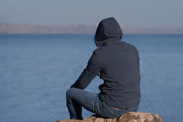Active European man is sitting on the stone near the lake and looking ahead. He is wearing a hood on his head. - 251879338