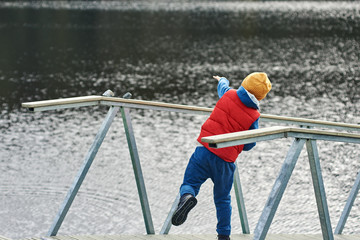 Boy is standing on the wooden quay and throwing stones to the lake.