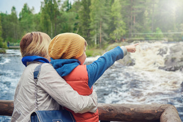 Young beautiful European woman and her son are standing on the wooden bridge and enjoying the picturesque view of the wild rough river. Boy is pointing out somewhere on the other side of the river.