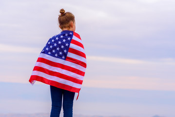 Young little girl holding American flag on amazing sky, mountain and meadow nature background at sunset. Memorial Day, Independence Day federal holidays in the United States.