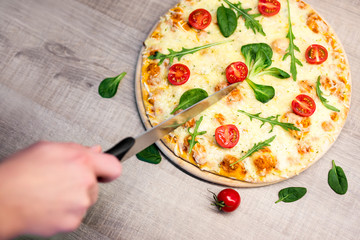 close up of male hand with big knife cutting pizza with tomatoes and herbs over wooden table