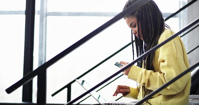 Good looking african woman with long dreadlocks using her credit card to buy something online using her notebook she sitting on the modern stairs in the office building