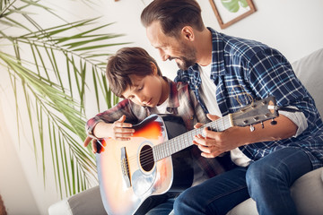 Father and little son at home sitting on sofa dad teaching boy playing guitar cheerful