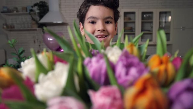 Closeup adorable mixed race son proudly holding gorgeous bouquet of multicolored tulips as gift for dear mom at Women's day. Portrait of smiling little boy offering beautiful flowers to camera