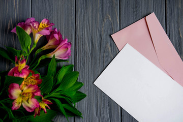 Mock up blank paper on a dark wooden background with natural flowers of pink color. Blank, frame for text. Greeting card design with flowers. Aalstroemeria on wooden background. View from above