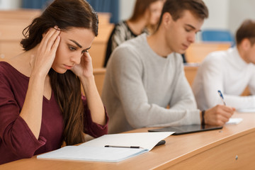 Serious female student sitting at table in lecture hall, keeping hands near head and thinking over difficult test. Concentrated girl looking down at notebook at university. Concept of studying.