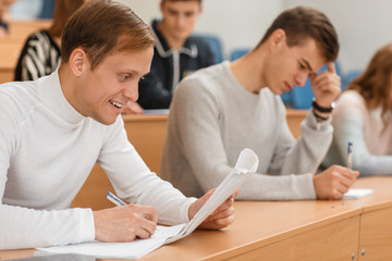 Cunning male student in white sweater sitting at table, smiling and copying notes at university. Cheerful man learning and getting high education. Concept of interesting studying.
