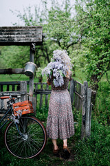 blonde girl in a dress is standing with her back and holding a bouquet of lilac near the bicycle on the background of a wooden well