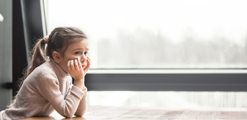 Charming little girl sitting at a wooden table by the window.