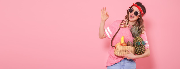 Beautiful young girl in pink t-shirt and glasses, holds a full straw bag of fruit on pink background. Shows hand OK