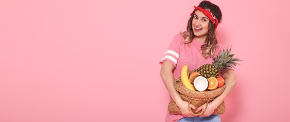 Beautiful young girl in pink t-shirt, holds a full straw bag of fruit on pink background