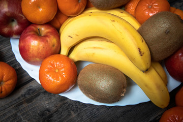 Fresh fruits on a white plate on old wooden table. Bananas, kiwi, apples and tangerines.