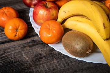 Fresh fruits on a white plate on old wooden table. Bananas, kiwi, apples and tangerines.
