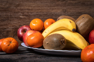 Fresh fruits on a white plate on old wooden table. Bananas, kiwi, apples and tangerines.