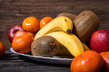 Naklejka na ściany i meble Fresh fruits on a white plate on old wooden table. Bananas, kiwi, apples and tangerines.