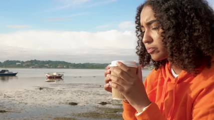 Panning shot of a beautiful mixed race African American girl teenager young woman wearing orange hoodie, drinking takeout coffee by a harbor looking sad or thoughtful
