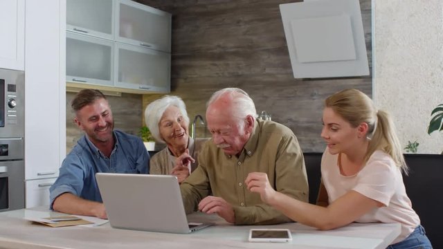Medium shot of aged couple with help of their adult children mastering laptop while sitting at table in kitchen