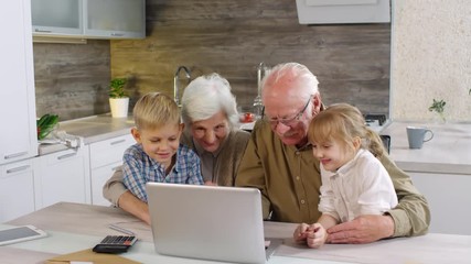 Medium shot of old couple sitting at kitchen table in front of computer, then their grandchildren coming to them, and they starting browsing together