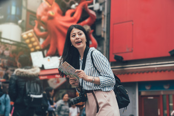 Fototapeta na wymiar vintage style photo of smiling gril backpack photographer standing in front of takoyaki food vendor shop with huge octopus animal advertisement hanging on wall in background. lady cheerful with map