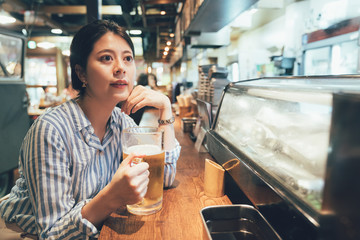 vintage style portrait of elegant woman holding ice cold beer glasses sitting at bar counter alone after work. asian office lady enjoy alcohol in japanese pub. young people drinks izakaya nightlife