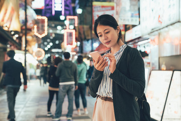 vintage style of young japanese woman texting sms cellphone in city at night. lady backpacker standing on street with lights in shinsekai with tsutenkaku tower in back. girl touch mobile phone screen