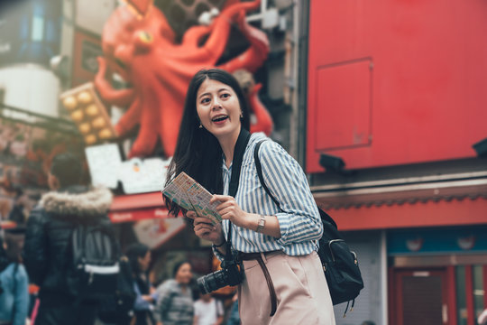 vintage style photo of smiling gril backpack photographer standing in front of takoyaki food vendor shop with huge octopus animal advertisement hanging on wall in background. lady cheerful with map