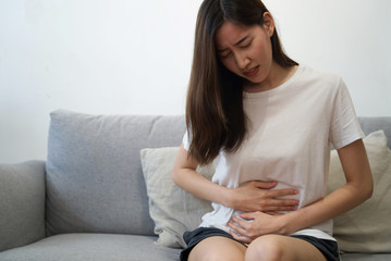 Fototapeta premium Young beautiful Asian girl is feeling unwell sitting on sofa. She is having stomach ache problem because of menstruation and she is holding her belly by both hands. Sickness and healthcare concept.
