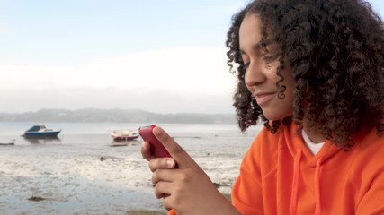Beautiful mixed race African American girl teenager young woman wearing orange hoodie, sitting on a sea front using her smart cell phone for social media or texting