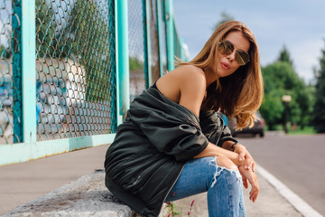 Fototapeta na wymiar Fashion portrait of trendy young woman wearing sunglasses, jeans with halls and bomber jacket in the city