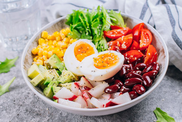 Close-up of Healthy vegan lunch bowl. Vegetables salad with avocado, egg, red bean, tomato, radish, corn, green leaves. Concrete background.